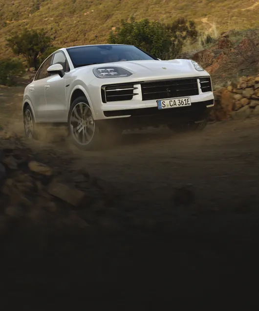 Porsche Cayenne E-Hybrid Coupé in Carraraweissmetallic fährt über Schotterstraße durchs Gelände. Sicht auf Front / Bug.