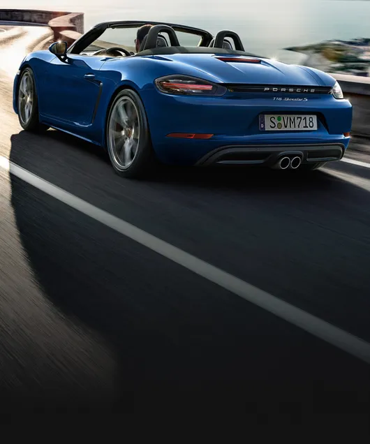 Porsche 718 Boxster S en Bleu Gentiane métallisé roulant sur une route côtière. Vue arrière et latérale.