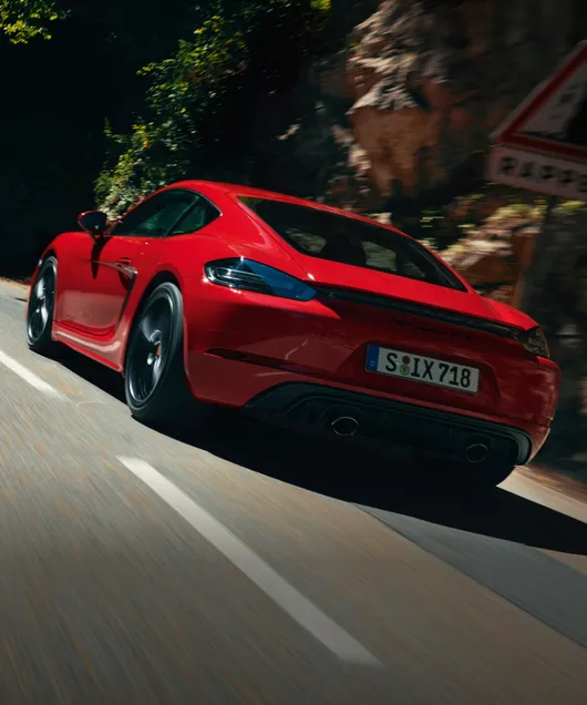 Porsche 718 Cayman GTS 4.0 en color Rojo Carmín circulando por una carretera. Vista de la parte trasera con ópticas traseras oscurecidas.