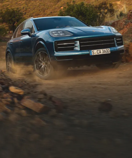 Porsche Cayenne em Azul Montego metálico passando por uma estrada de cascalho. Vista dianteira/spoiler.