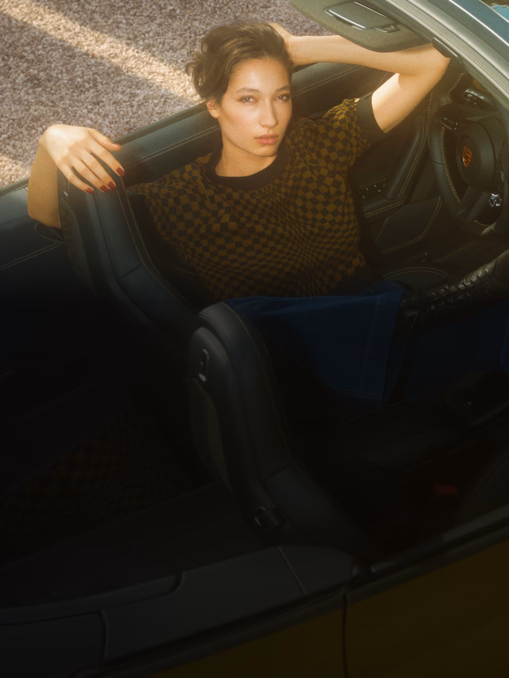 A woman wearing the Spirit 70 collection by Porsche Lifestyle casually sitting sideways in the driver seat of a Porsche Cabriolet.