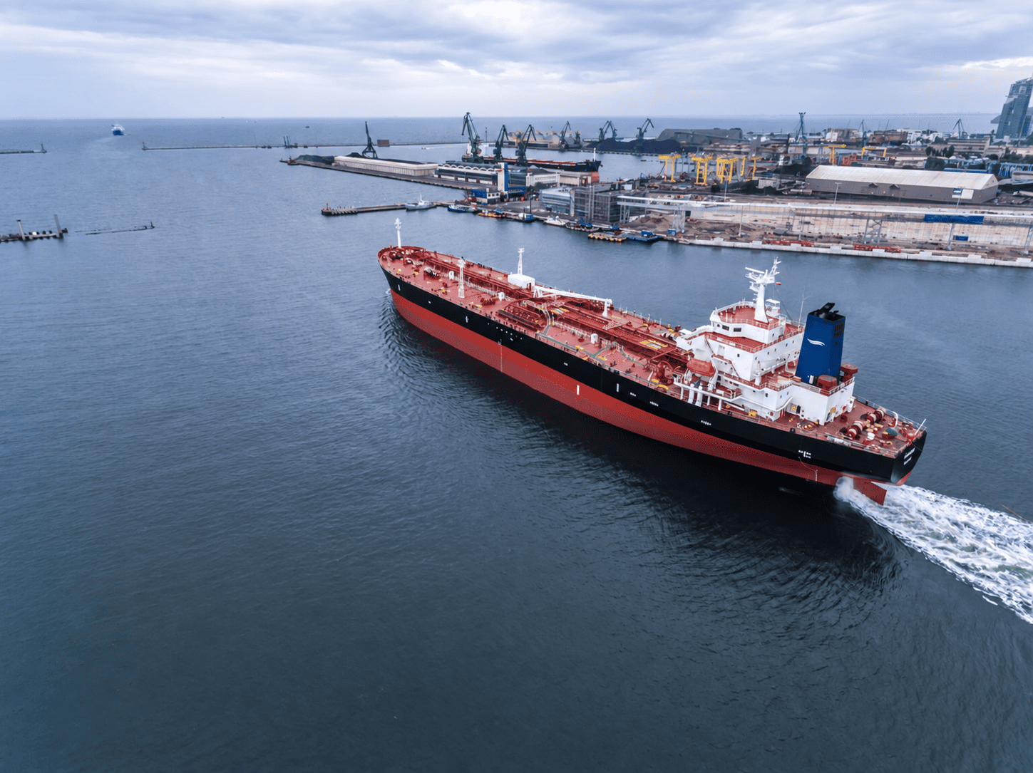 Large cargo ship navigating through a port with cranes and industrial buildings in the background under a cloudy sky.