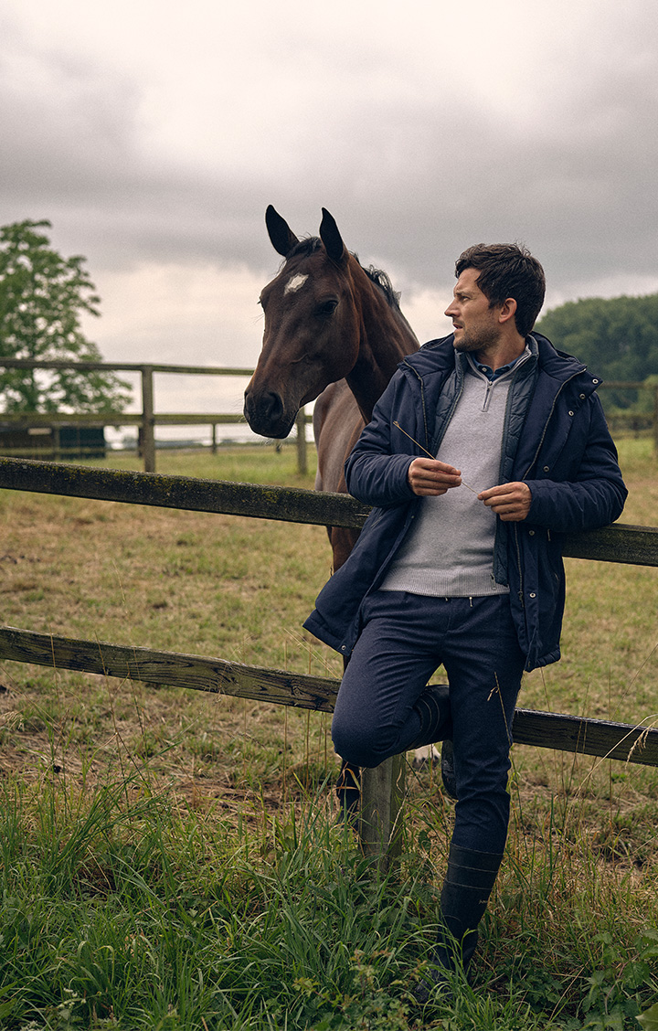 Model leunt tegen hek, achter hem staat een paard