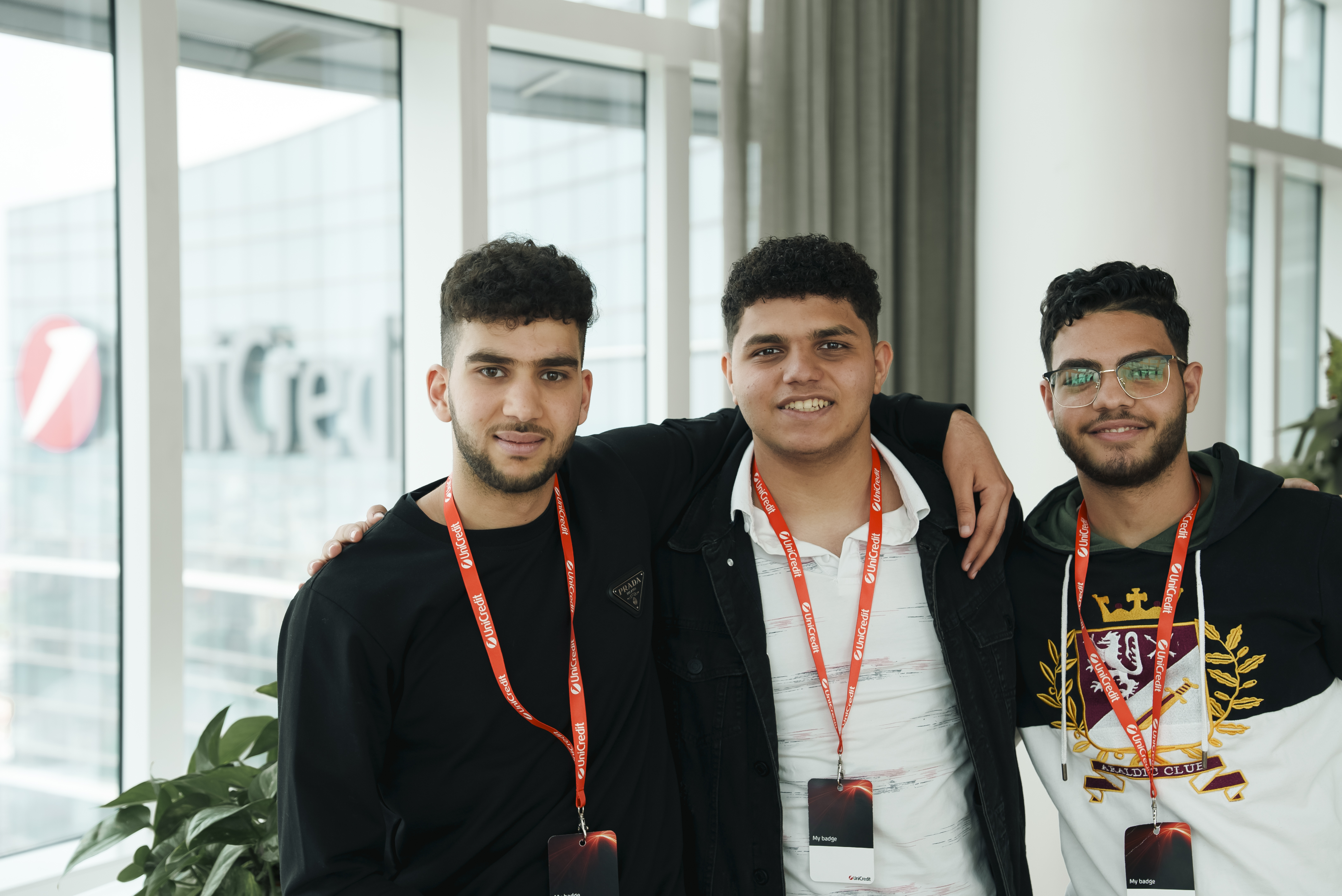 Tre uomini sorridenti con il distintivo della conferenza posano insieme al chiuso, vicino a grandi finestre.