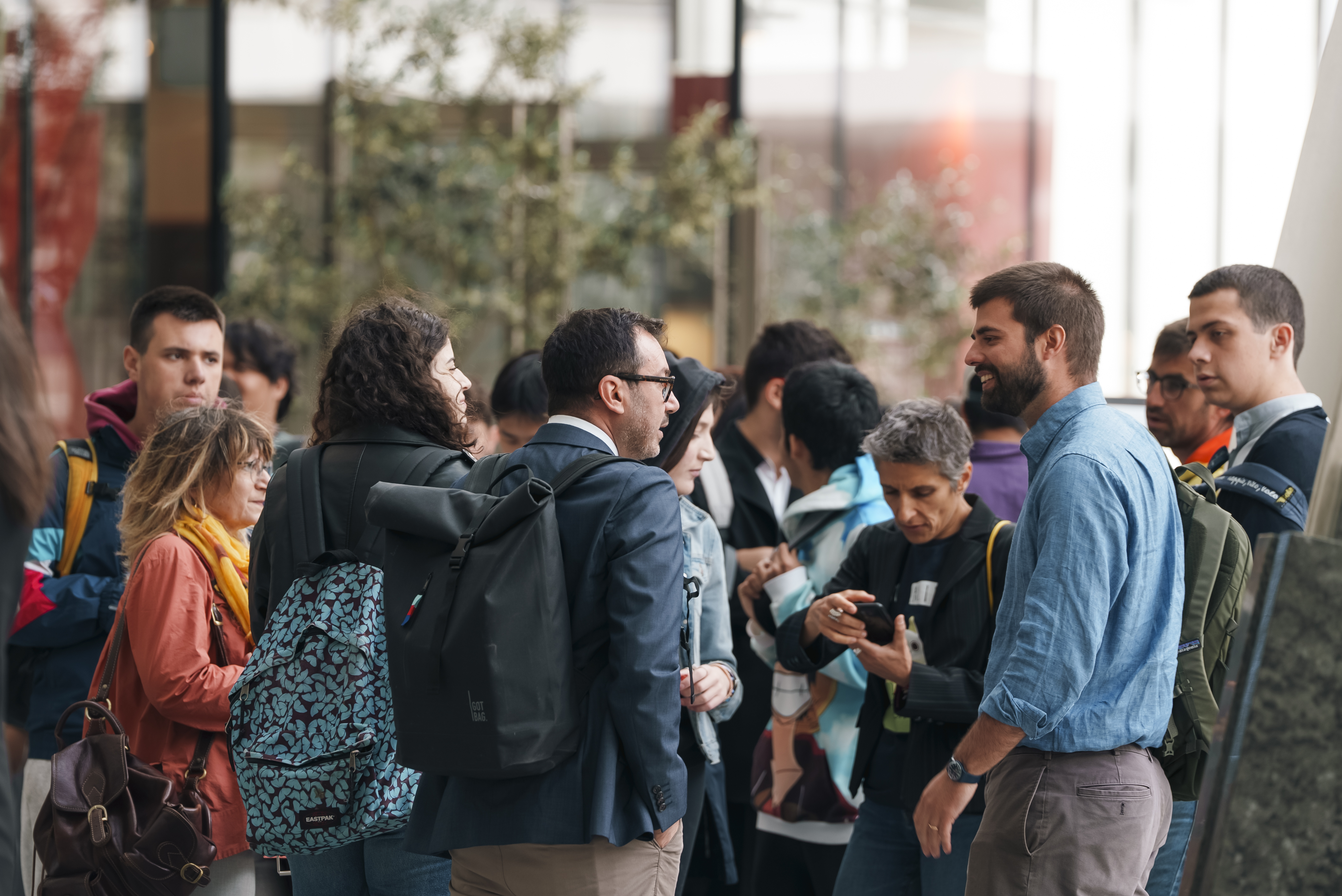 A group of people engaged in conversation, some with backpacks, in an indoor setting with plants in the background.