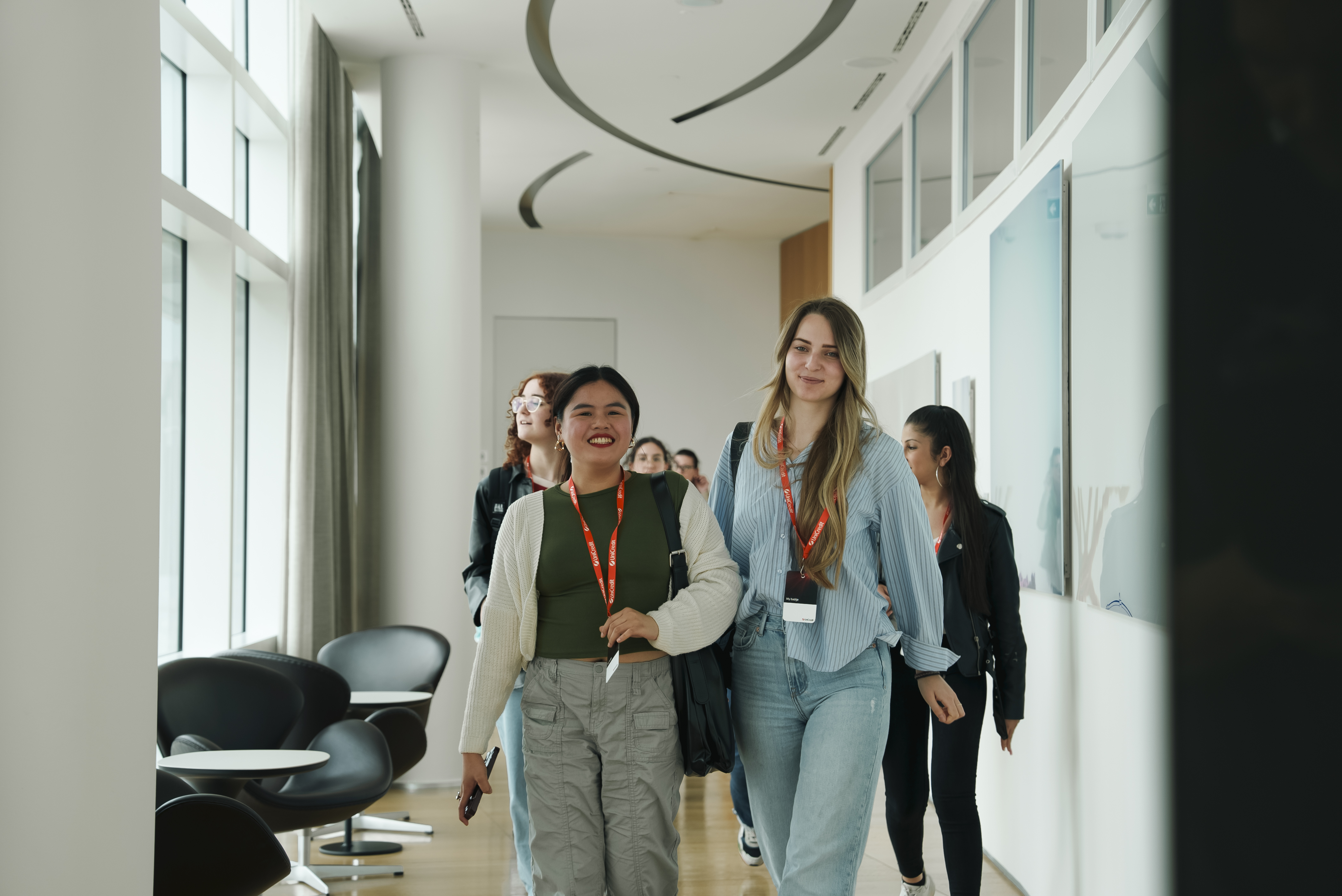 A group of people wearing lanyards walking down a bright hallway with large windows and modern chairs.