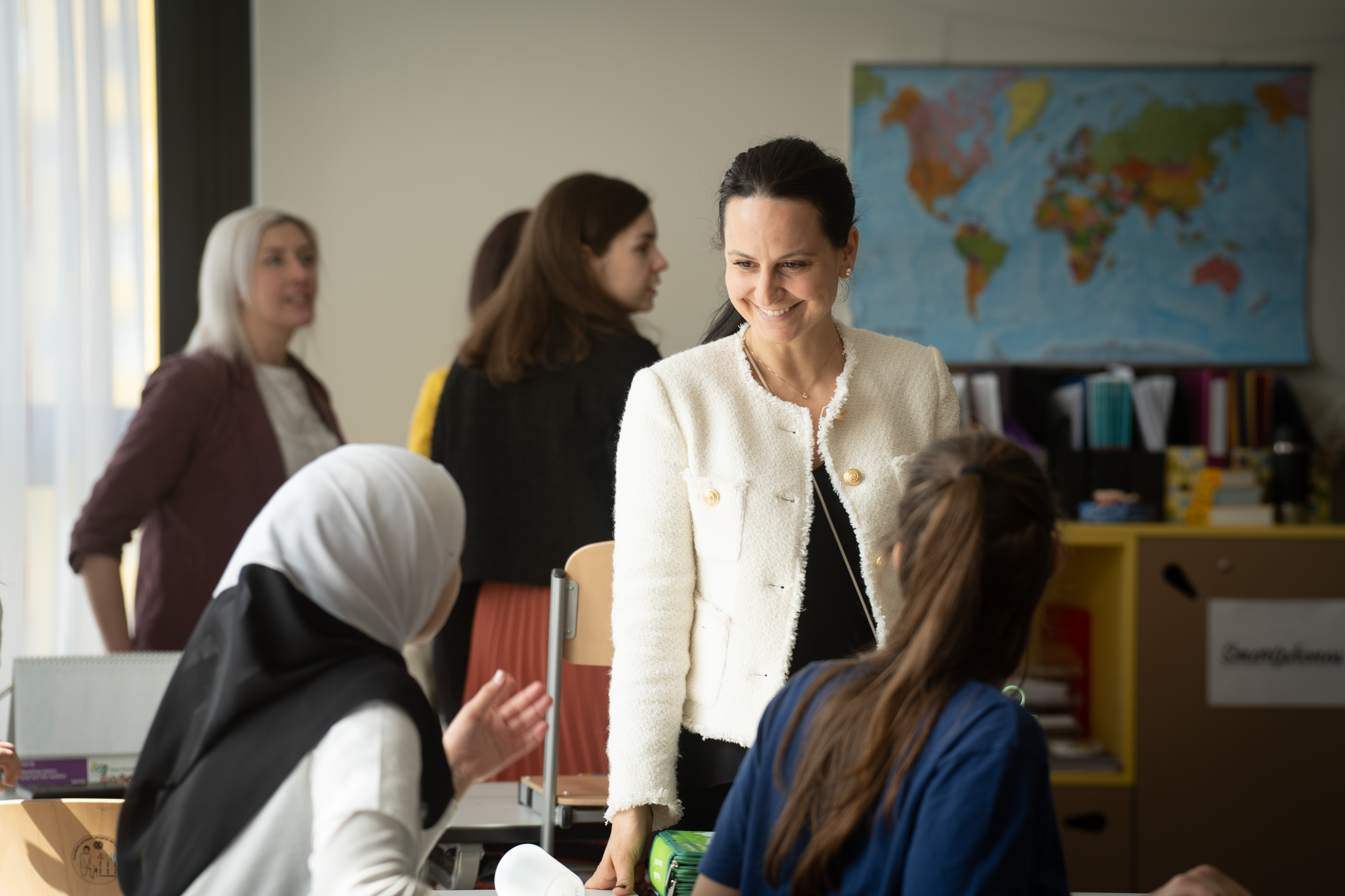 Una donna con un blazer bianco sorride mentre interagisce con due donne sedute in un'aula. Sullo sfondo una mappa del mondo e una libreria.