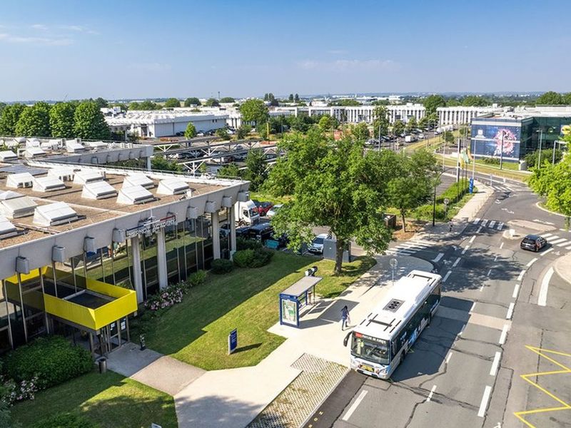 Aerial view of a modern industrial park with buildings, trees, roads, a bus, and cars under a clear blue sky.