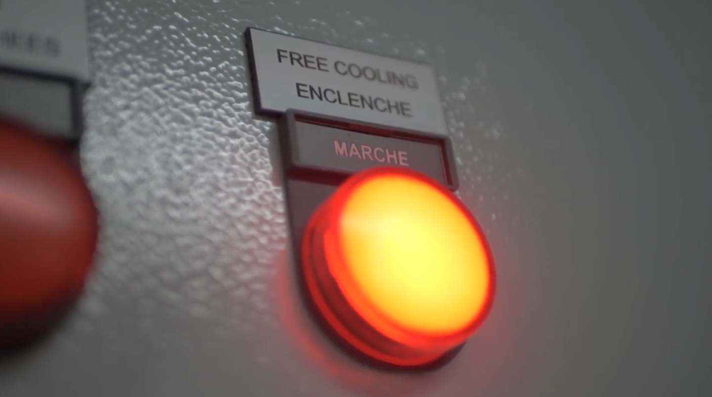 Close-up view of a control panel featuring a bright red illuminated button, with labels in French. The labels include 'FREE COOLING,' 'ENCLENCHE' (Engage), and 'MARCHE' (On), indicating a system for managing cooling operations.