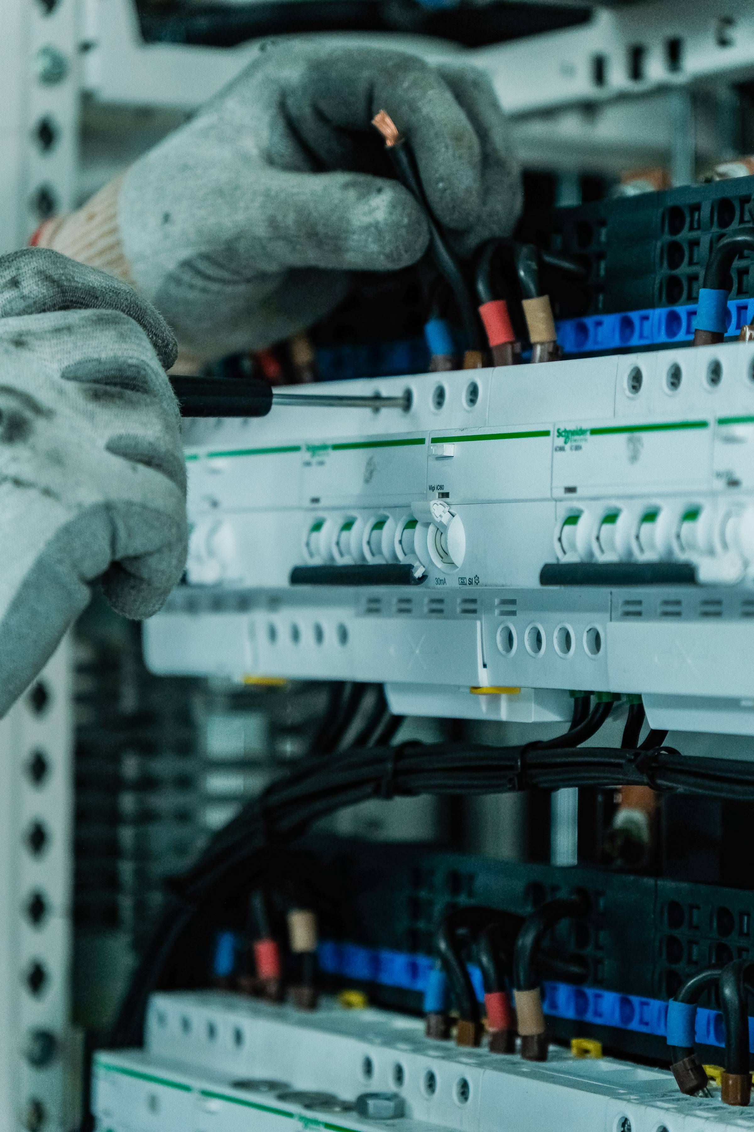 Electrician wearing gloves adjusts wiring in a circuit breaker panel, focusing on connections and switches in a detailed electrical setup.
