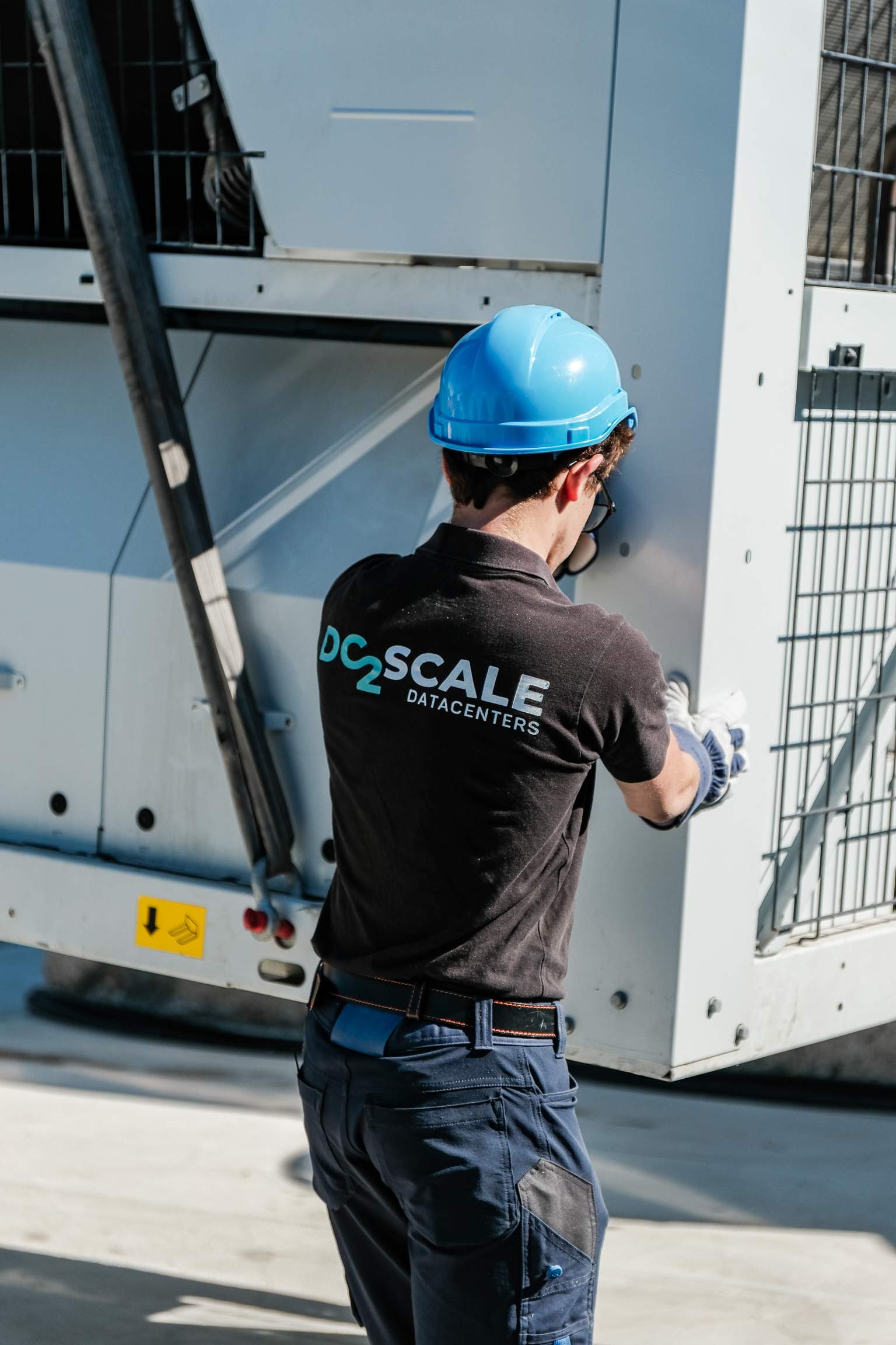 A worker in a blue hard hat and gloves adjusts equipment outdoors, wearing a "DC2Scale Datacenters" shirt.
