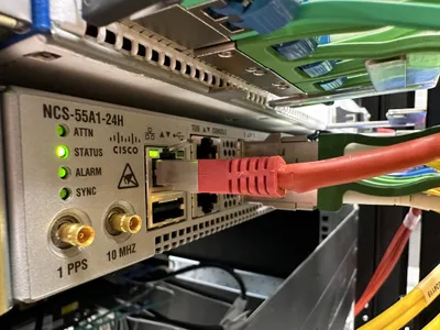 Close-up of a Cisco NCS-55A1-24H network device with LED indicators labeled ATT, STATUS, ALARM, and SYNC. A red Ethernet cable is connected.