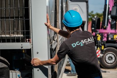 A worker in a blue hard hat and "DC2Scale Datacenters" shirt pushes a large metal panel outdoors, with machinery visible in the background.