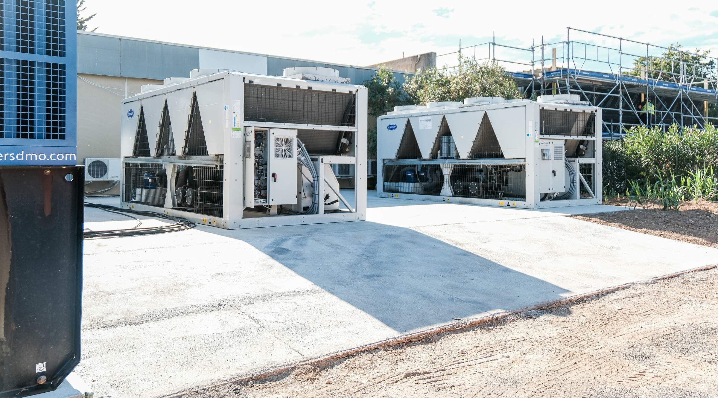Outdoor industrial air conditioning units on a concrete pad near a building, surrounded by scaffolding and greenery under a clear sky.