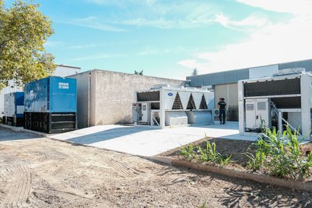 Outdoor view of industrial equipment on a sunny day, featuring large blue Kohler SDMO backup generators and white Carrier air conditioning or chiller units on concrete slabs in the DC2SCALE MRS1 Marseille Datacenter