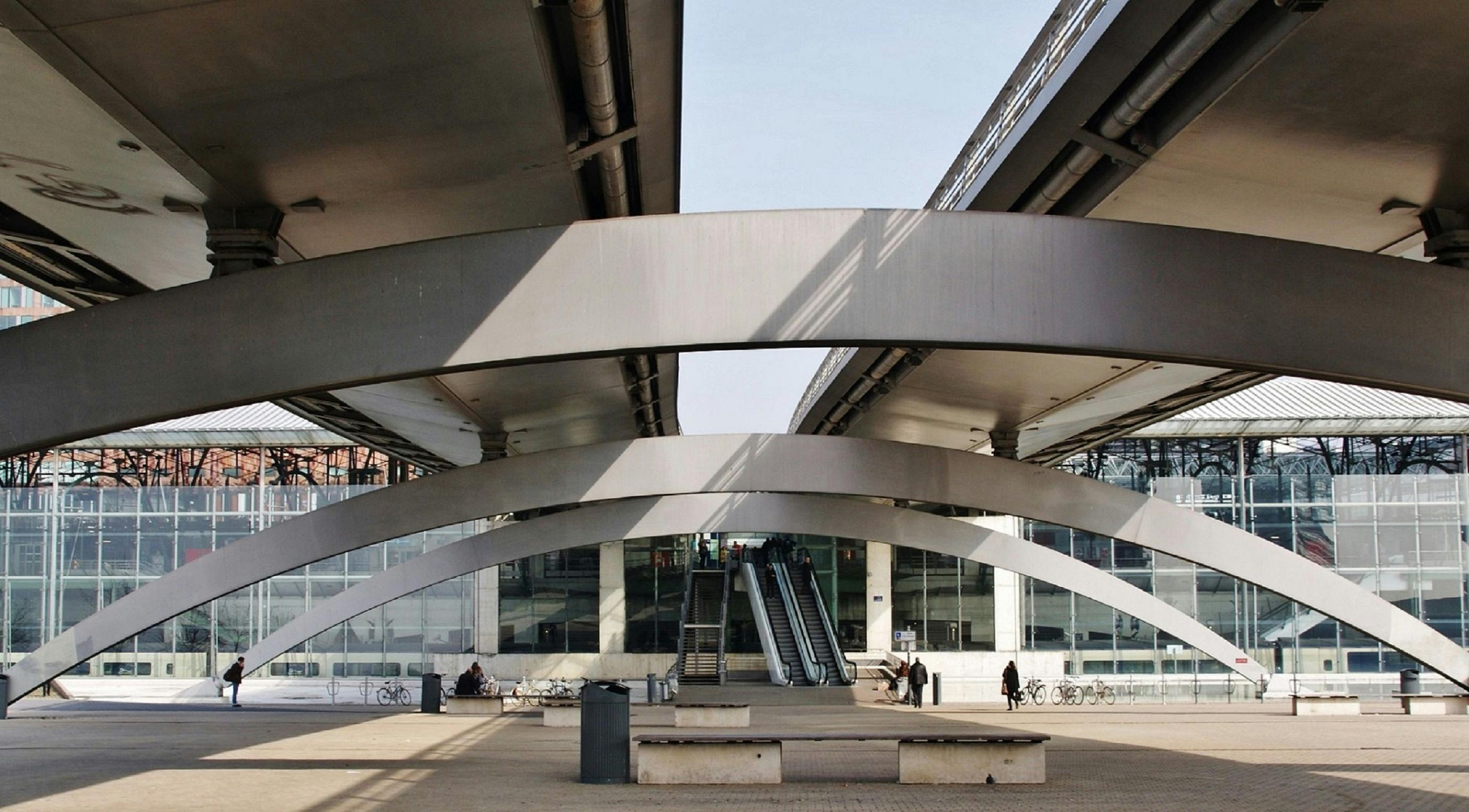 Modern architectural structure with large, curved metal beams and glass facade. People walk near escalators under the beams. In the city of Lille, France.
