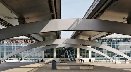 Modern architectural structure with large, curved metal beams and glass facade. People walk near escalators under the beams. In the city of Lille, France.