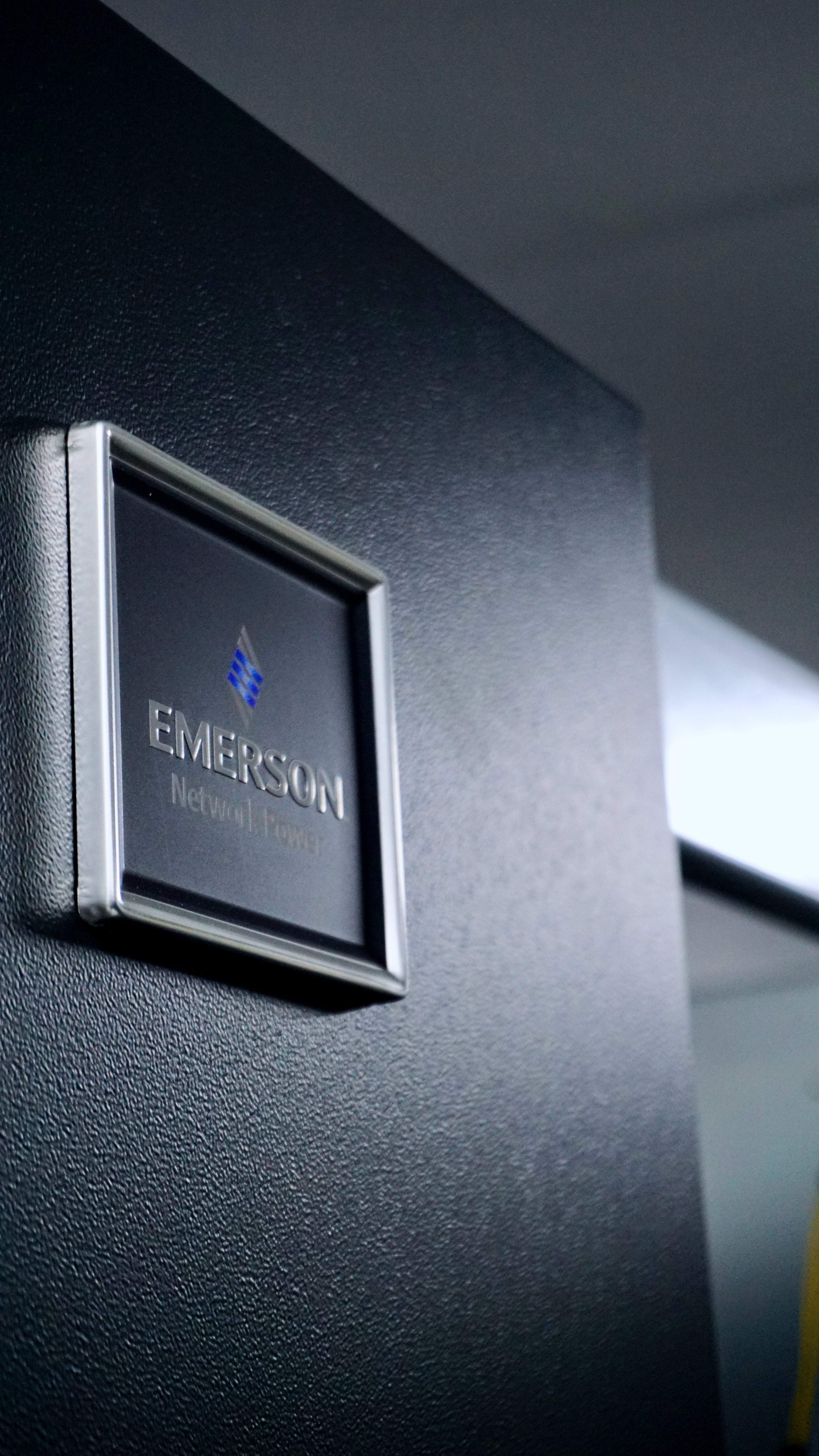 Close-up of a black Emerson Network Power equipment panel with a silver-framed logo, in a dimly lit environment.