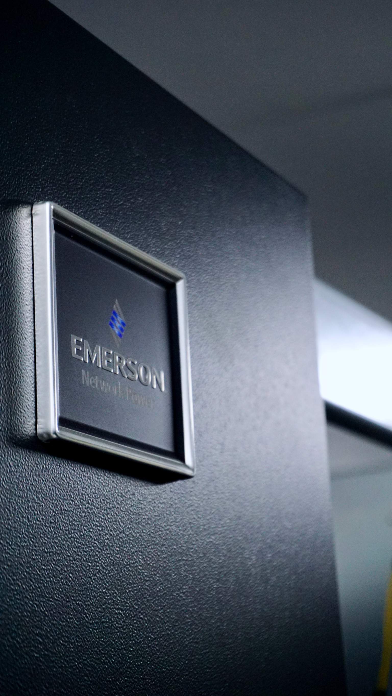Close-up of a black Emerson Network Power equipment panel with a silver-framed logo, in a dimly lit environment.