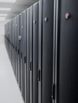 Black and grey server racks with an APC logo are aligned in a long corridor in a data center. The perspective is from a low angle. The focus is on the front racks and the rest of the corridor is blurry.