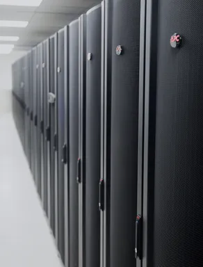 Black and grey server racks with an APC logo are aligned in a long corridor in a data center. The perspective is from a low angle. The focus is on the front racks and the rest of the corridor is blurry.