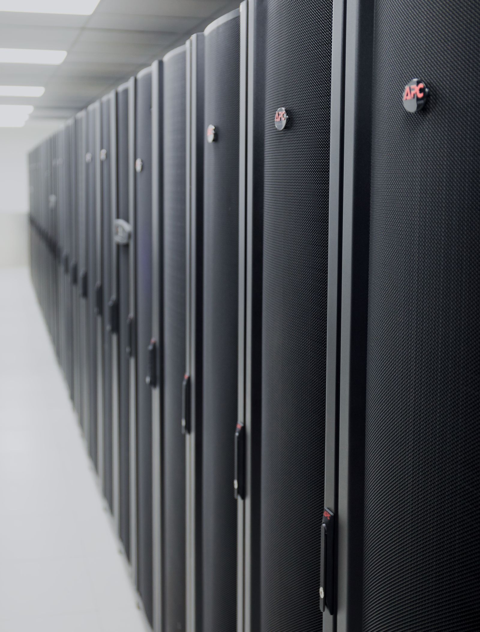 Black and grey server racks with an APC logo are aligned in a long corridor in a data center. The perspective is from a low angle. The focus is on the front racks and the rest of the corridor is blurry.