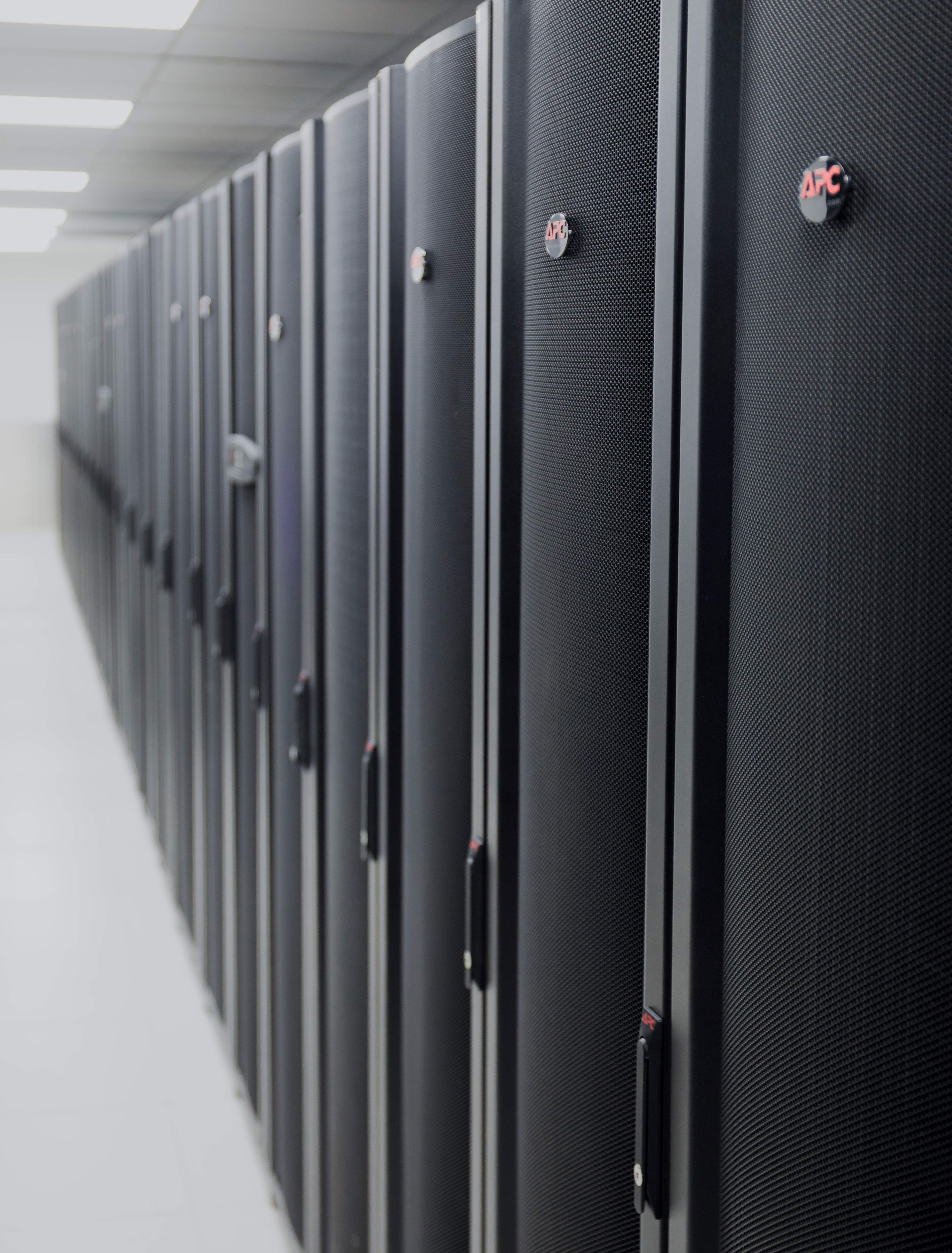 Black and grey server racks with an APC logo are aligned in a long corridor in a data center. The perspective is from a low angle. The focus is on the front racks and the rest of the corridor is blurry.