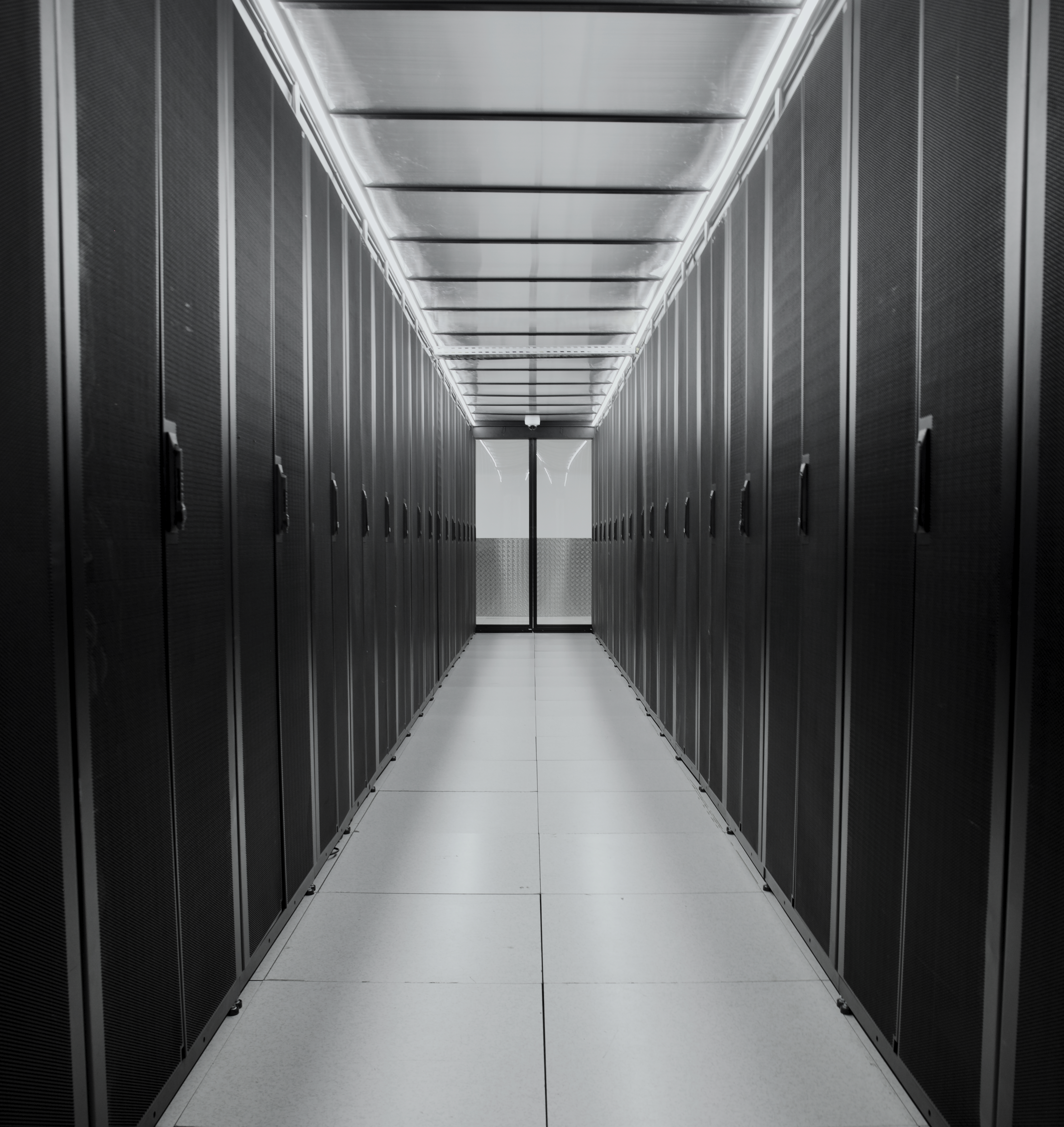 Black and white shot of a data center aisle. The symmetrical composition with the corridor in the middle emphasizes the long line of server racks on each side. The aisle is brightly lit by a luminous ceiling, contrasting with the dark racks.