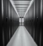 Black and white shot of a data center aisle. The symmetrical composition with the corridor in the middle emphasizes the long line of server racks on each side. The aisle is brightly lit by a luminous ceiling, contrasting with the dark racks.