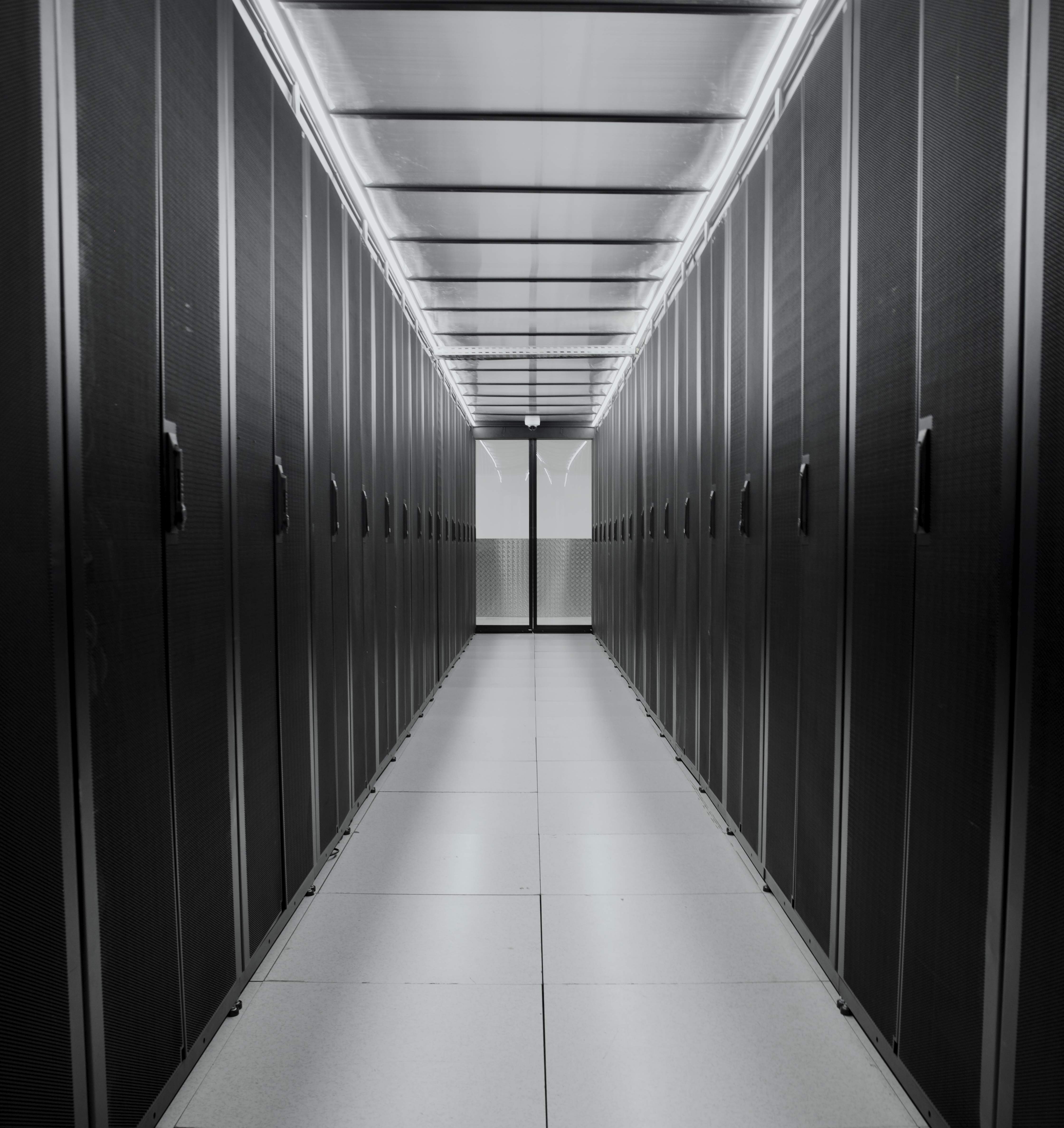 Black and white shot of a data center aisle. The symmetrical composition with the corridor in the middle emphasizes the long line of server racks on each side. The aisle is brightly lit by a luminous ceiling, contrasting with the dark racks.