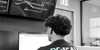 A technician from behind wearing a "DC2SCALE DATACENTER" t-shirt, working at a desk with a dual-monitor setup.