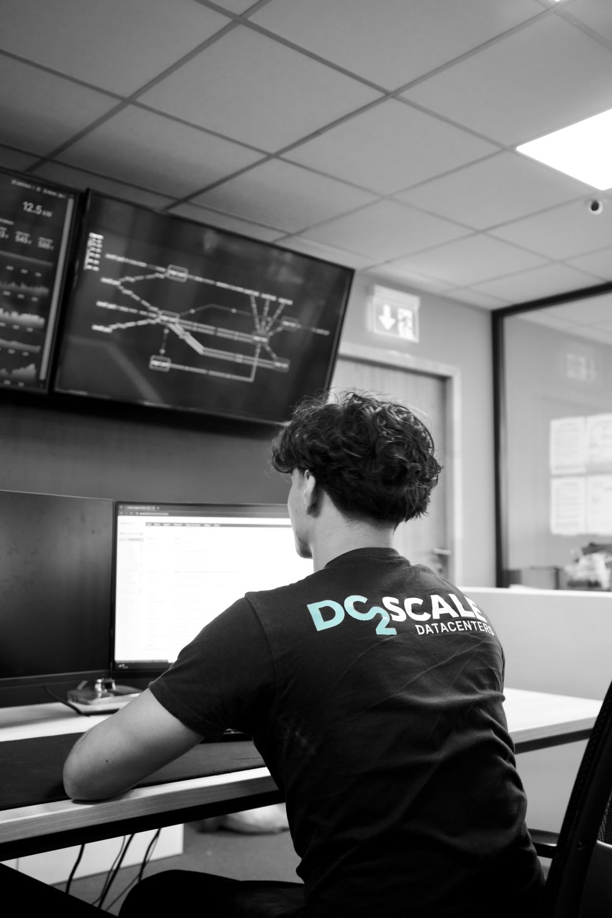 A technician from behind wearing a "DC2SCALE DATACENTER" t-shirt, working at a desk with a dual-monitor setup.