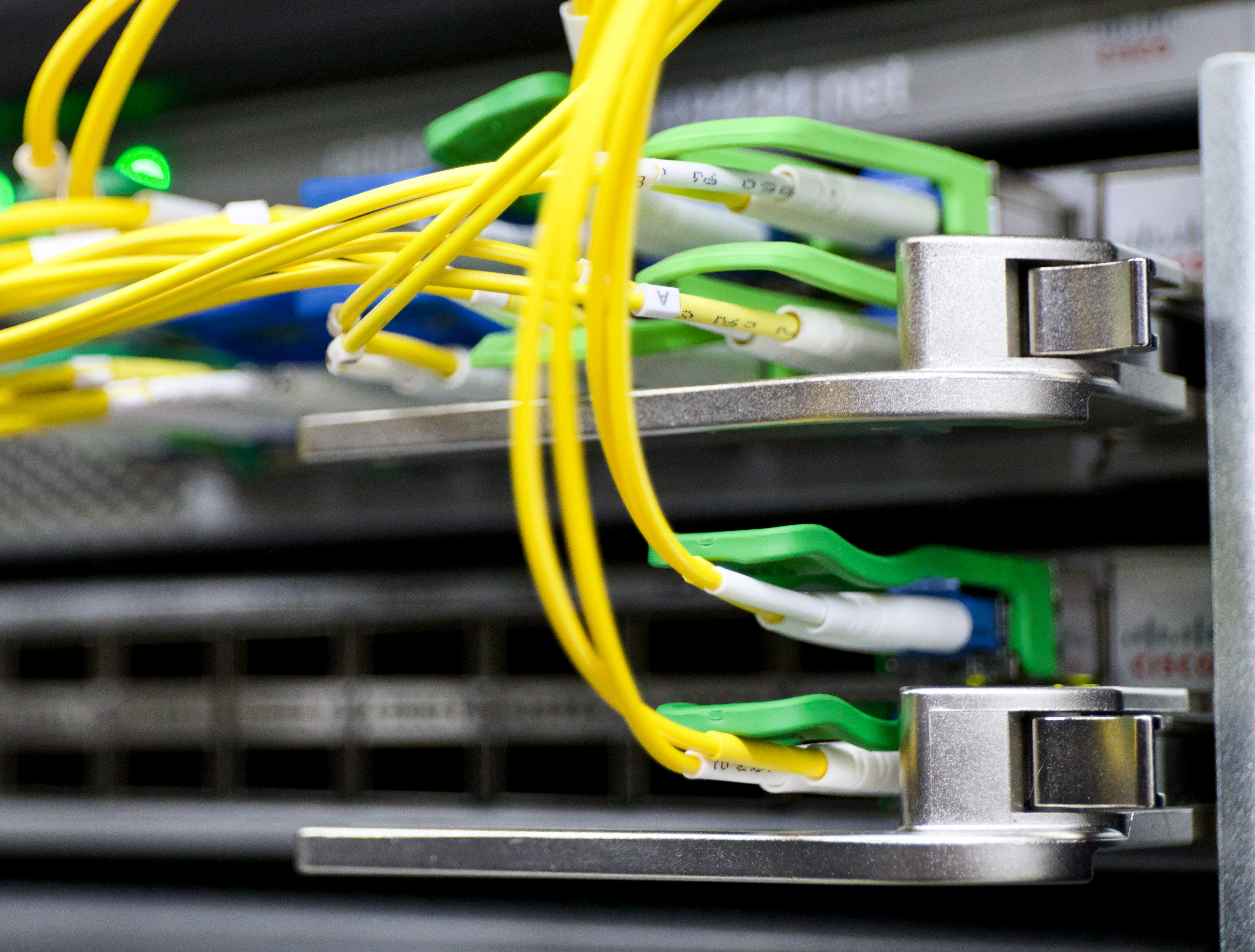 A close-up shot of yellow and green fiber cables plugged into a data center network equipment (Cisco). The cables are bright and colorful, standing out against the grey server. The background is out of focus.