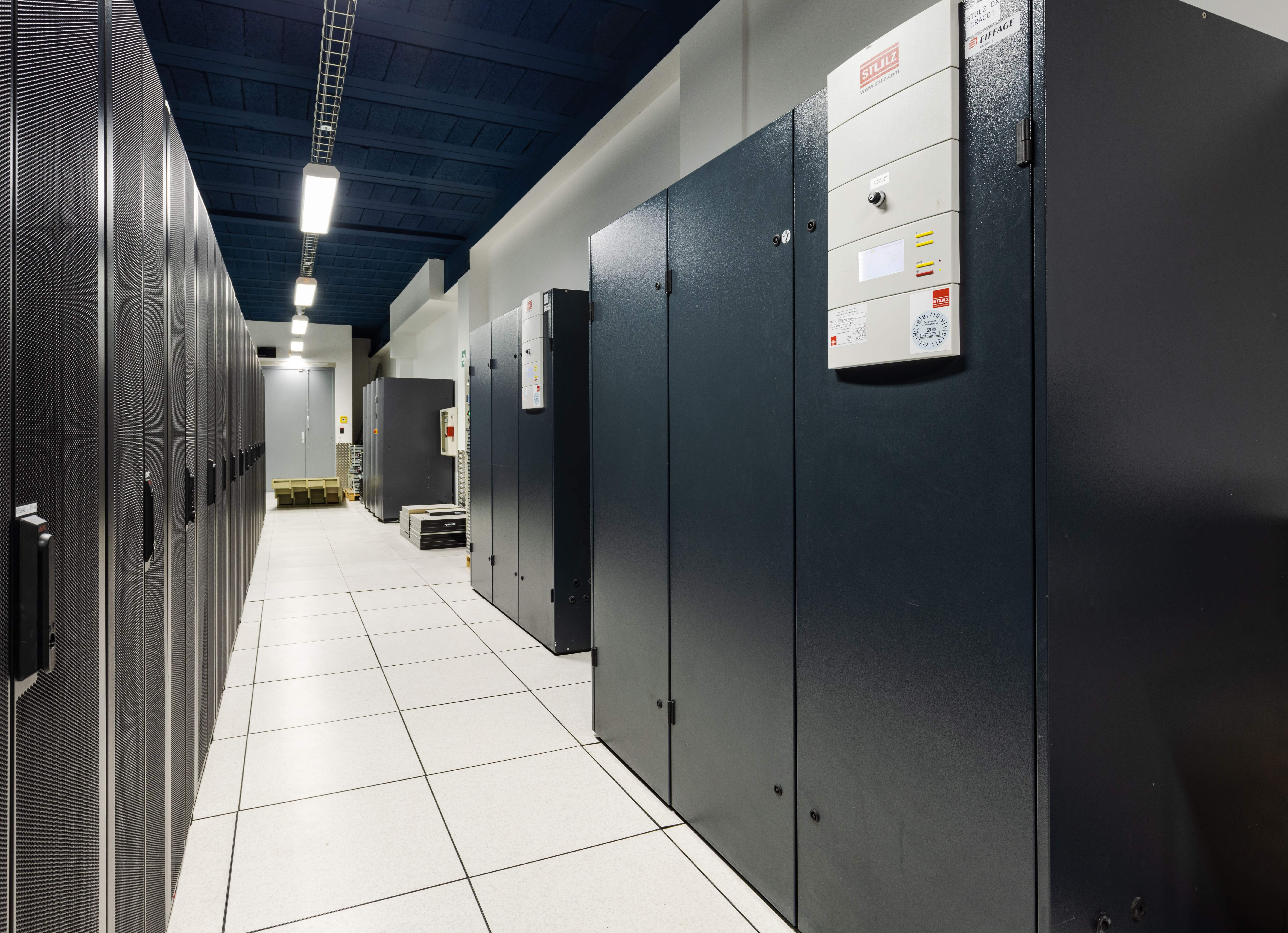 A wide-angle interior shot of a data center in Vélizy-Villacoublay, Île-de-France, France. On the left, rows of black server racks with perforated doors line the aisle. On the right, a series of large, dark grey Stulz Computer Room Air Conditioners (CRACs) are visible, providing essential cooling for the data center equipment. The floor is composed of white square tiles, and the ceiling is painted a distinctive dark blue, from which fluorescent lights are suspended, illuminating the entire space. The aisle is empty, highlighting the organized and functional environment of the facility.