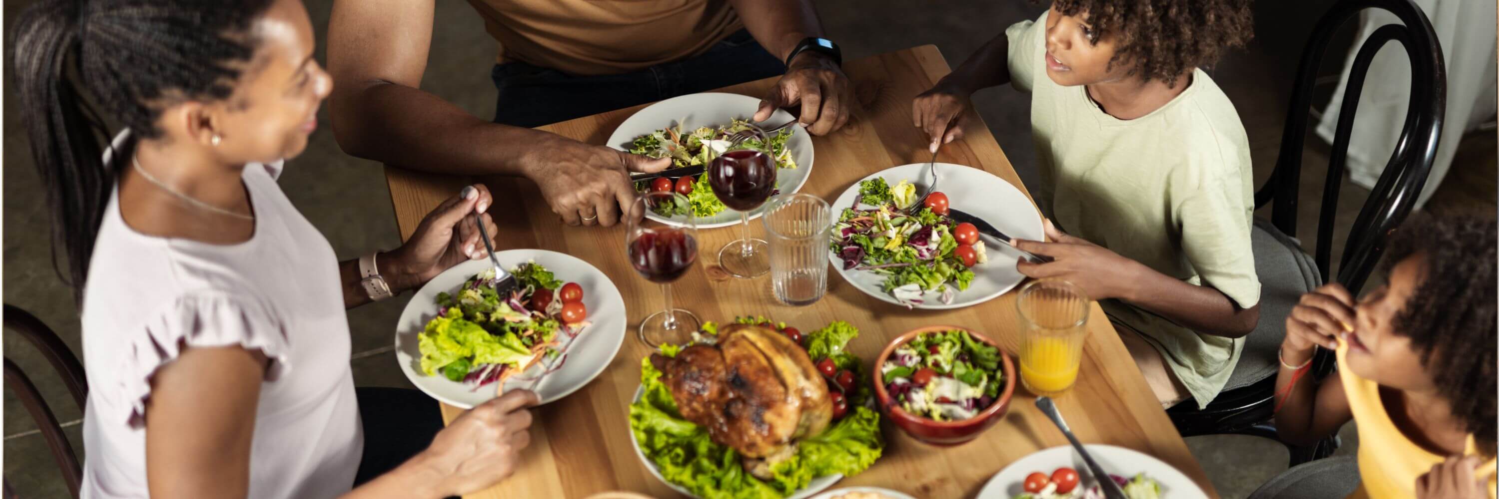 Family dining together, enjoying salads, roasted chicken, and drinks around a wooden table in a cozy setting.