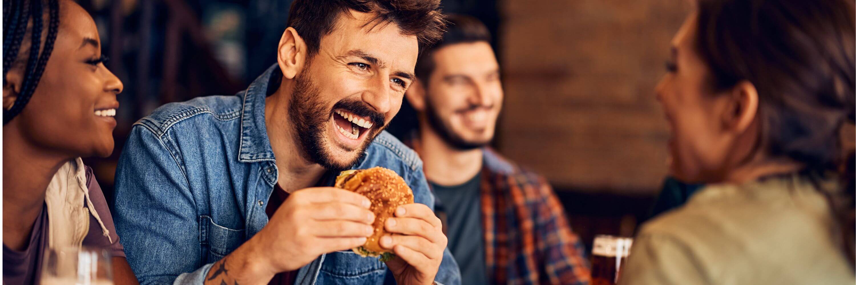 Friends laughing and enjoying burgers together at a restaurant, with one person holding a burger and others smiling warmly.