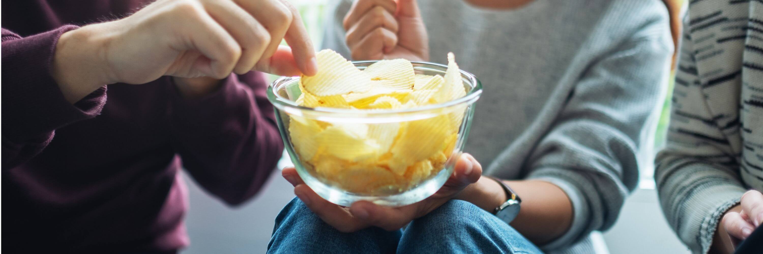Hands reaching for potato chips in a glass bowl, with people in sweaters sitting nearby.