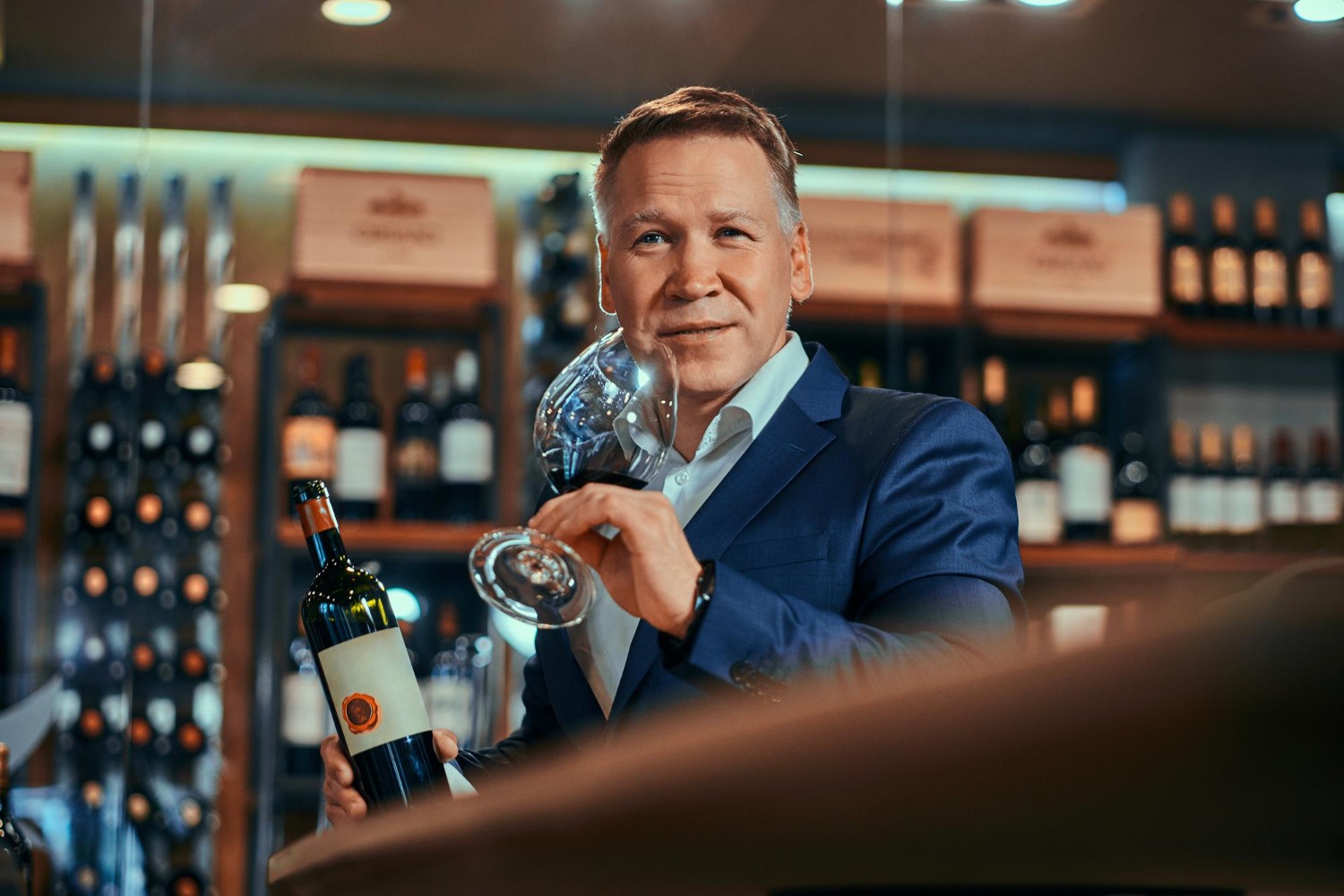 Man in a suit holding a wine bottle and glass, standing in a wine shop with shelves of bottles in the background.