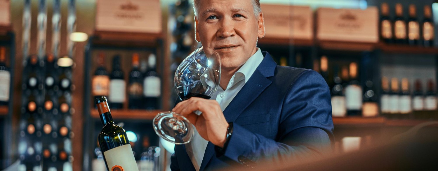 Man in a suit holding a wine bottle and glass, standing in a wine shop with shelves of bottles in the background.