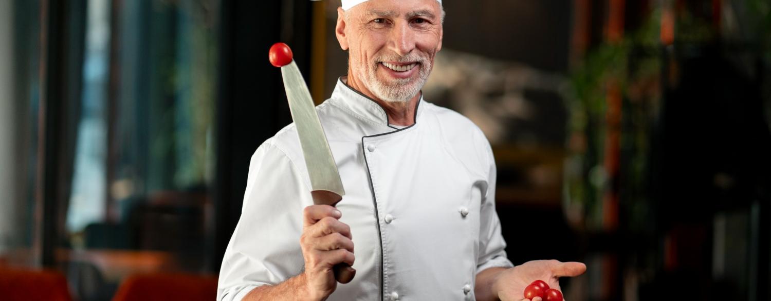 Smiling chef in a white uniform and hat holds a knife and cherry tomatoes in a restaurant kitchen.