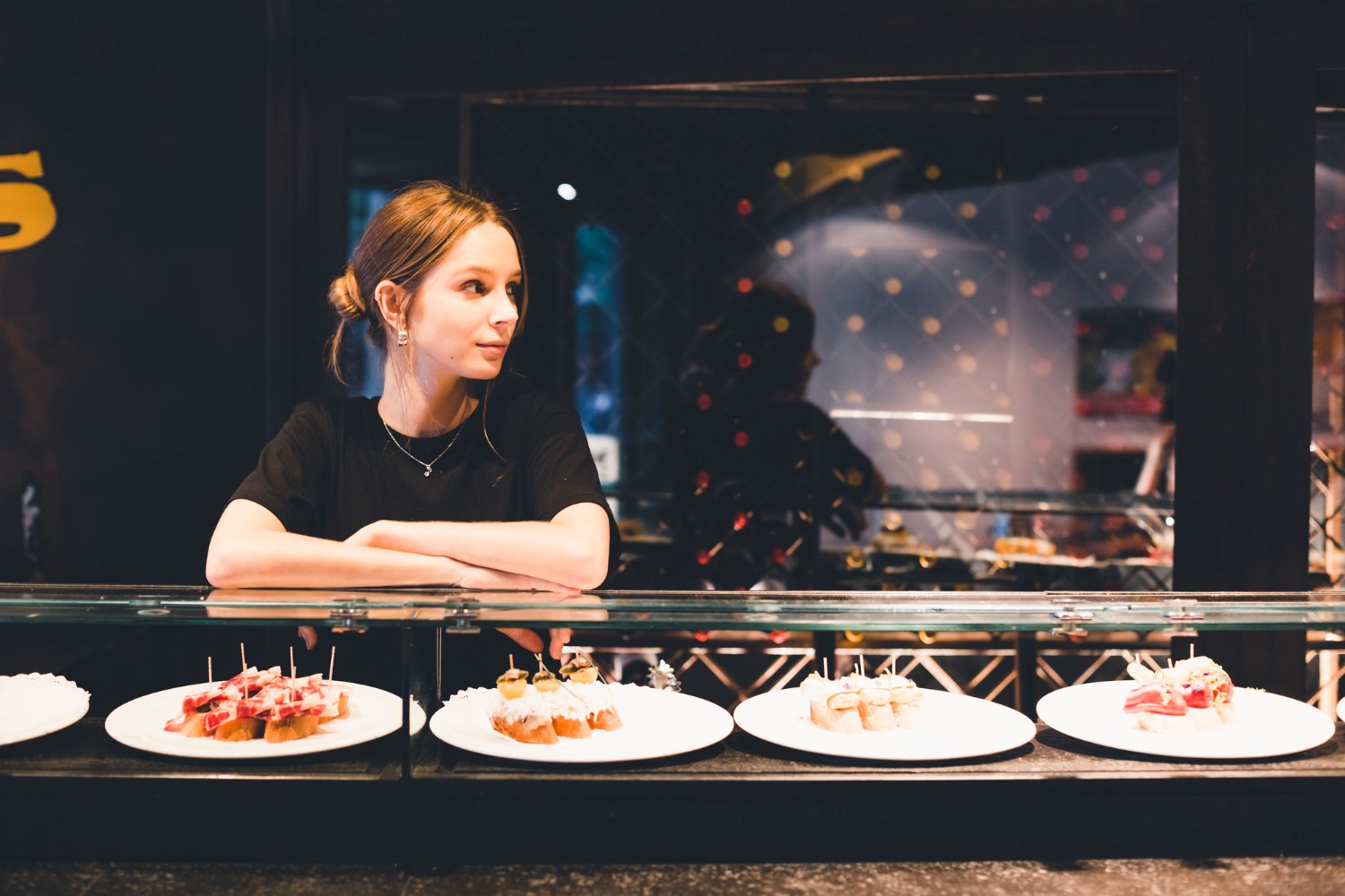 Woman standing behind a counter with plates of appetizers, looking to the side, in a dimly lit setting.