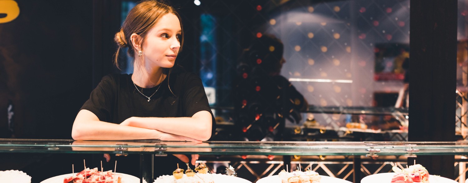 Woman standing behind a counter with plates of appetizers, looking to the side, in a dimly lit setting.