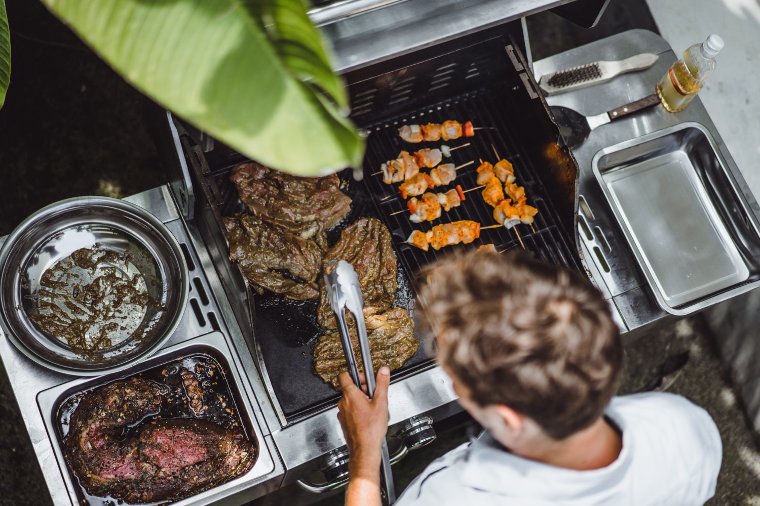 Person grilling meat and skewers on a barbecue, surrounded by cooking tools and dishes, under a leafy plant.