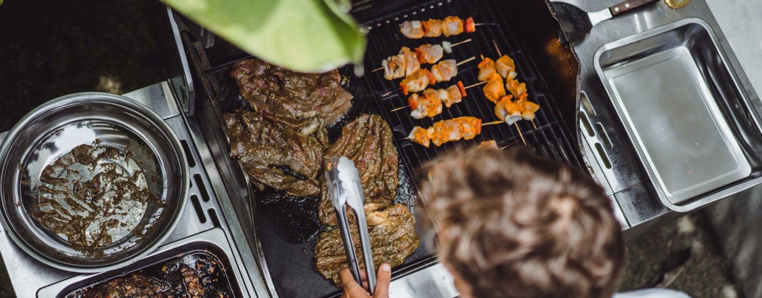 Person grilling meat and skewers on a barbecue, surrounded by cooking tools and dishes, under a leafy plant.