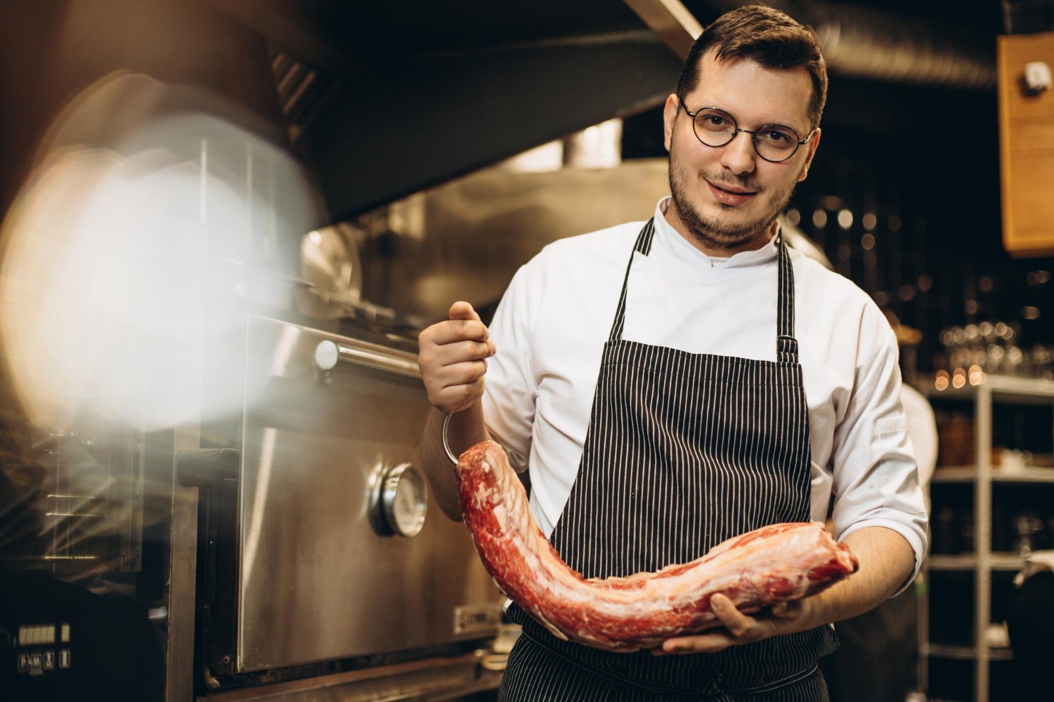 Chef in a kitchen holding a large cut of raw meat, wearing glasses, a white shirt, and a striped apron, standing near an oven.