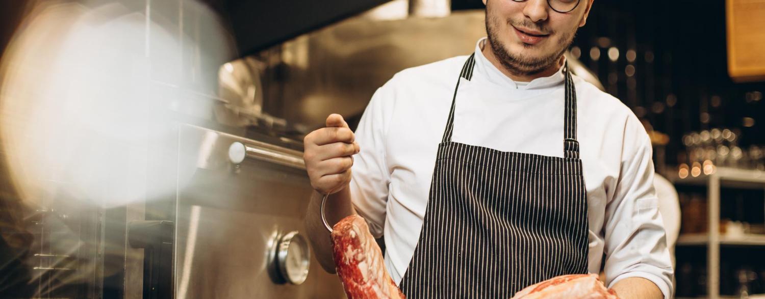 Chef in a kitchen holding a large cut of raw meat, wearing glasses, a white shirt, and a striped apron, standing near an oven.