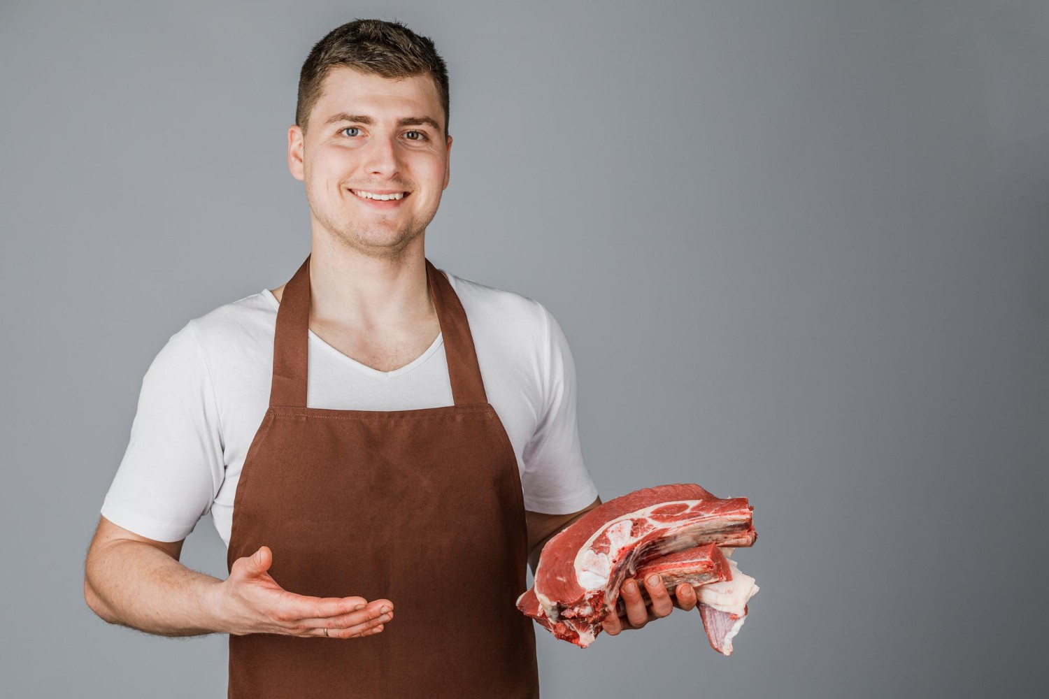 Smiling butcher in a brown apron holds raw meat cuts, gesturing with one hand against a gray background.