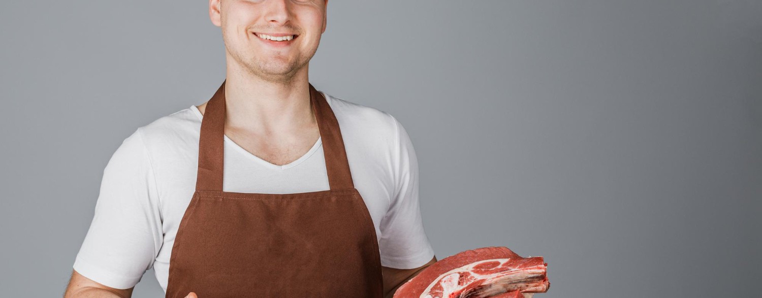 Smiling butcher in a brown apron holds raw meat cuts, gesturing with one hand against a gray background.