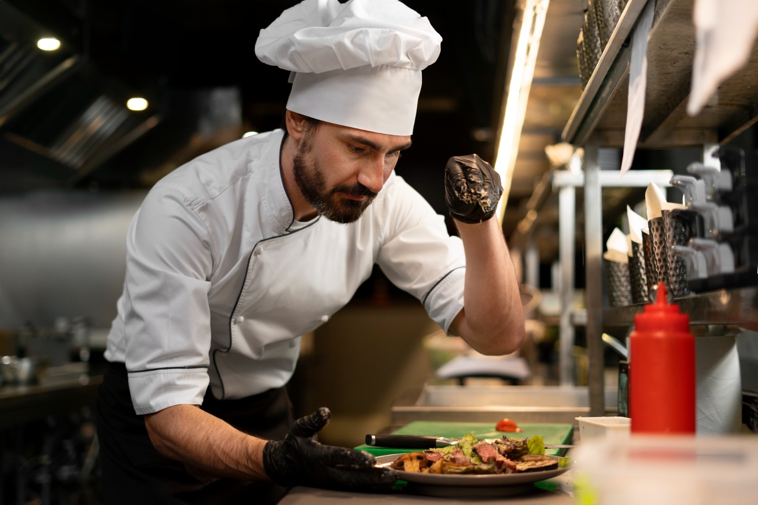Chef in a white uniform and hat garnishing a dish in a restaurant kitchen, with focused expression and gloved hands.