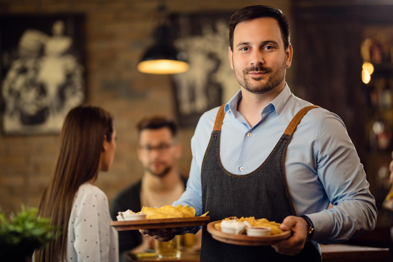 A waiter in a blue shirt and apron holds plates of food, standing in a cozy restaurant with two seated patrons in the background.
